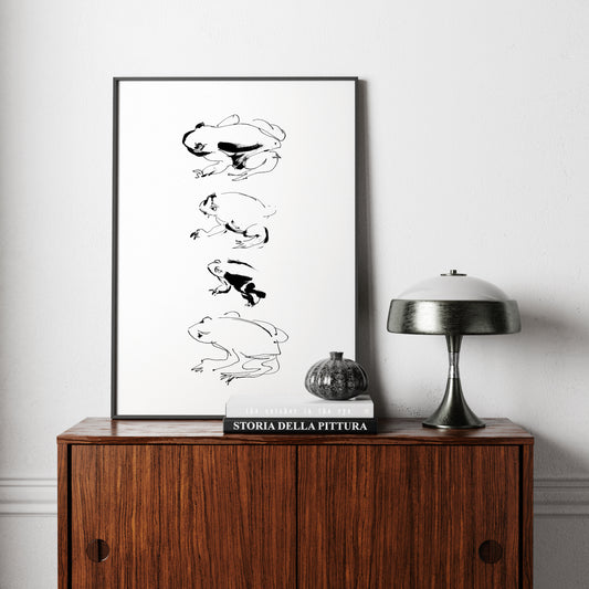 Framed black and white artwork of a frog study in ink on a wooden cabinet with a lamp and book.