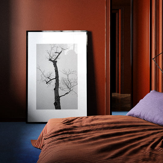 Framed artwork of a bare tree against a gray sky in a bedroom with red walls and a bed.