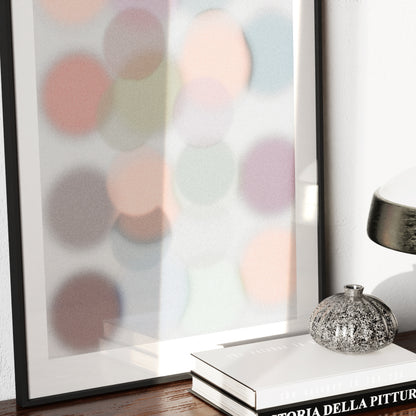 Detail of a framed abstract art print with pastel hue colors dots, with a lamp and books on a table in the foreground.