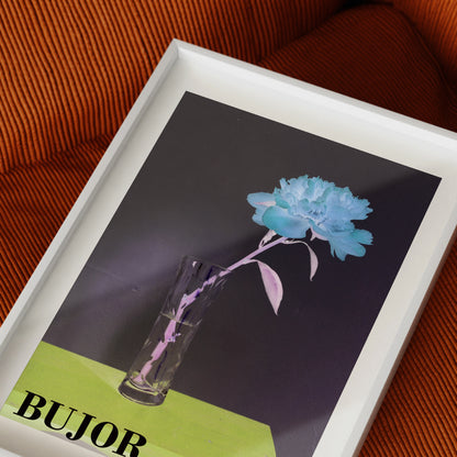 Framed artwork with a blue flower in a glass vase on a dark background, placed on an orange textured sofa surface.