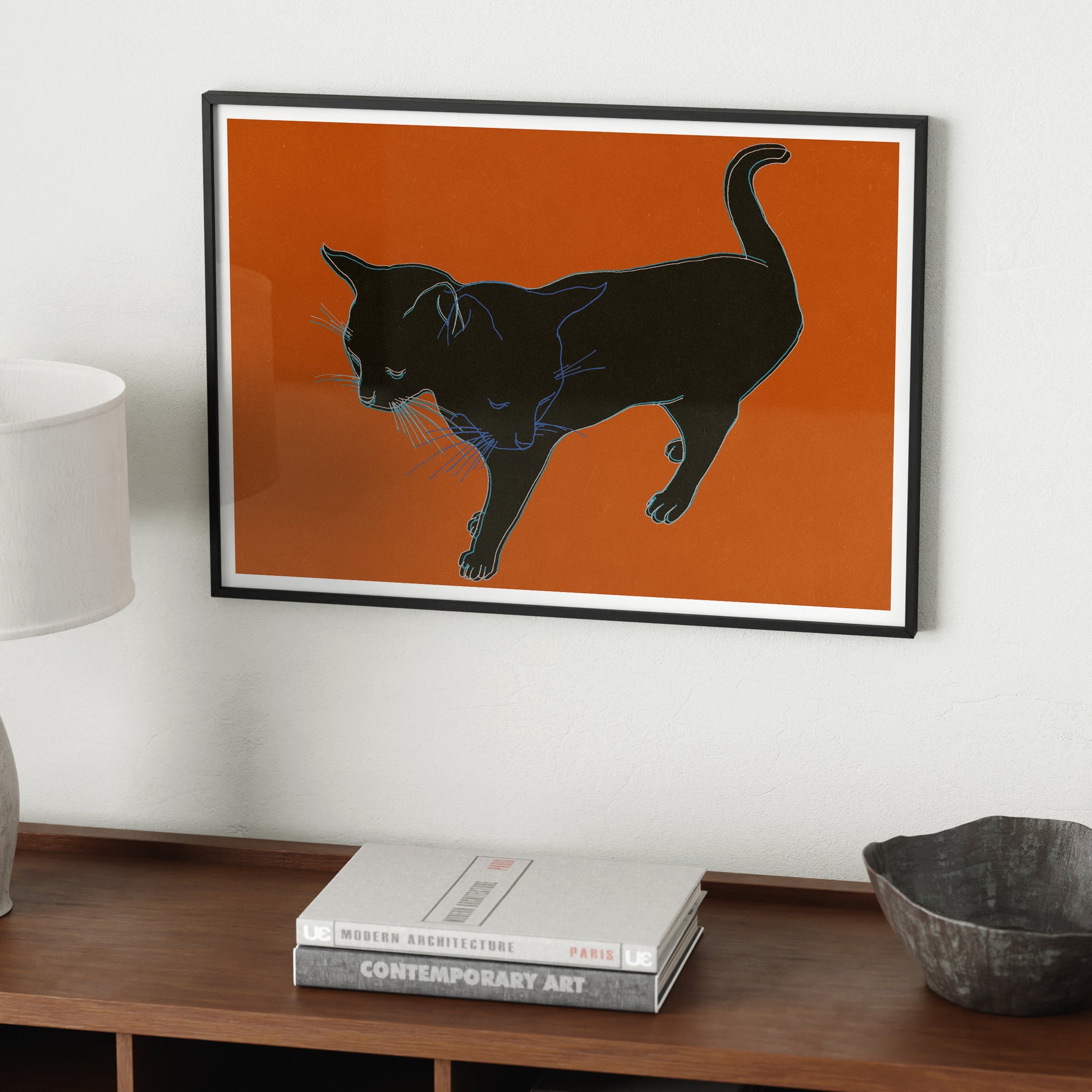 Framed art print with a black cat on an orange background hanging above a wood sideboard.