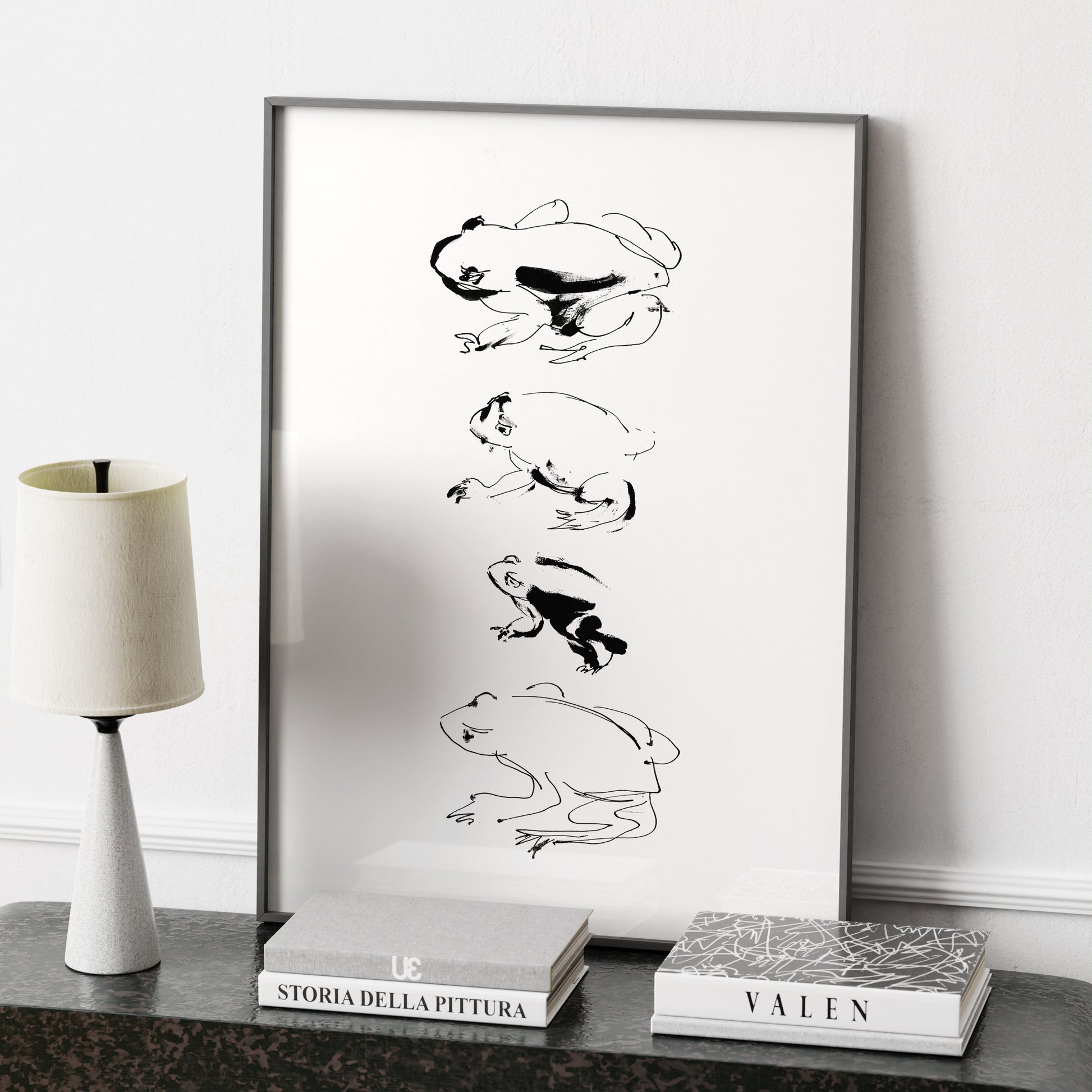 Framed black and white artwork of a frog study on a shelf with books and a lamp.