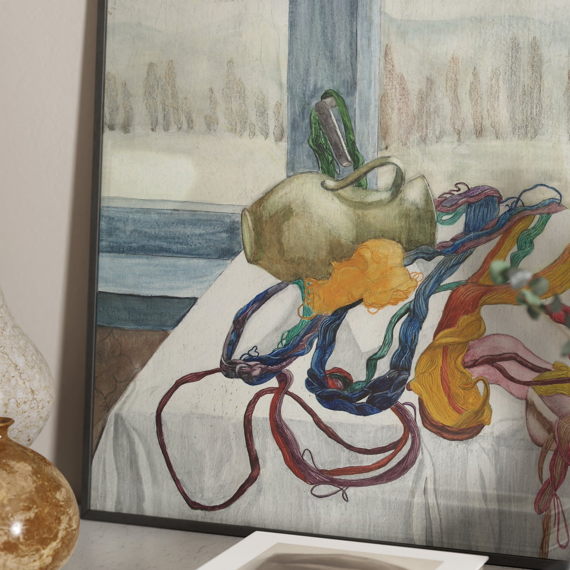 Detail of a figurative framed art print with colored threads arranged on a table, with a window in the background and a winter landscape.