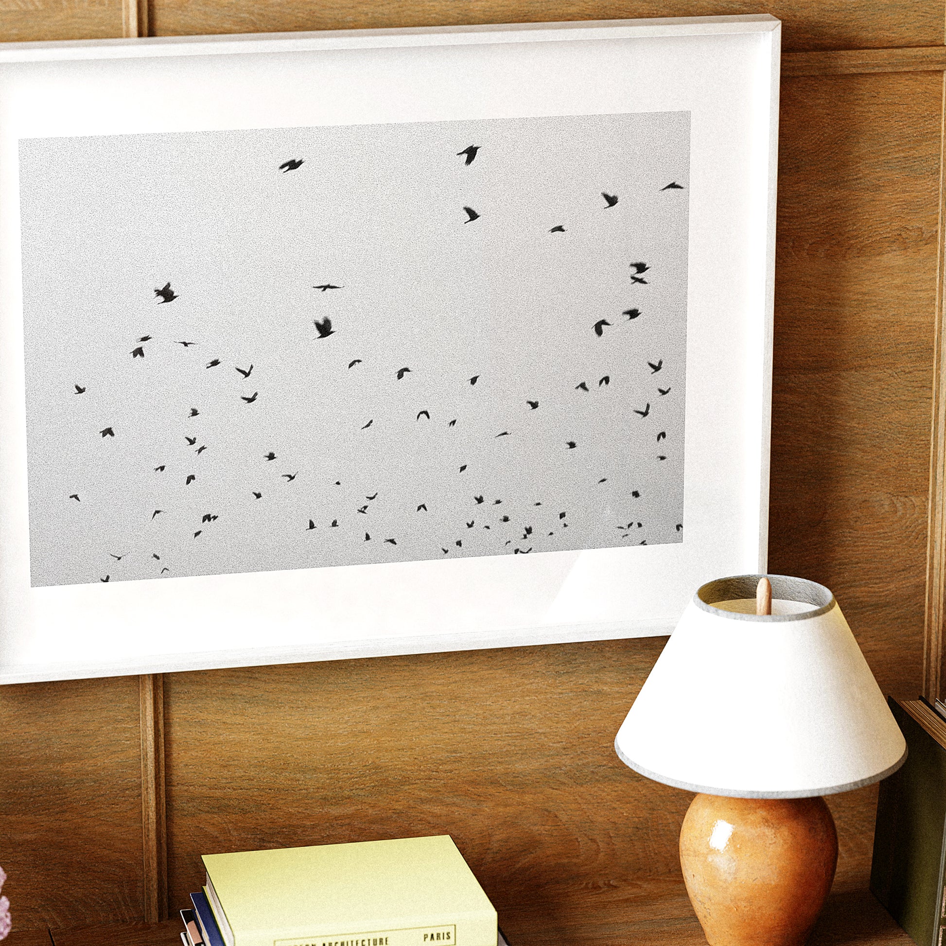 Framed artwork with black birds on a white background, placed on a wooden surface next to a lamp.