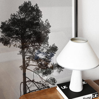 White lamp on a side-table with a book and a black and white photography print with a tree.