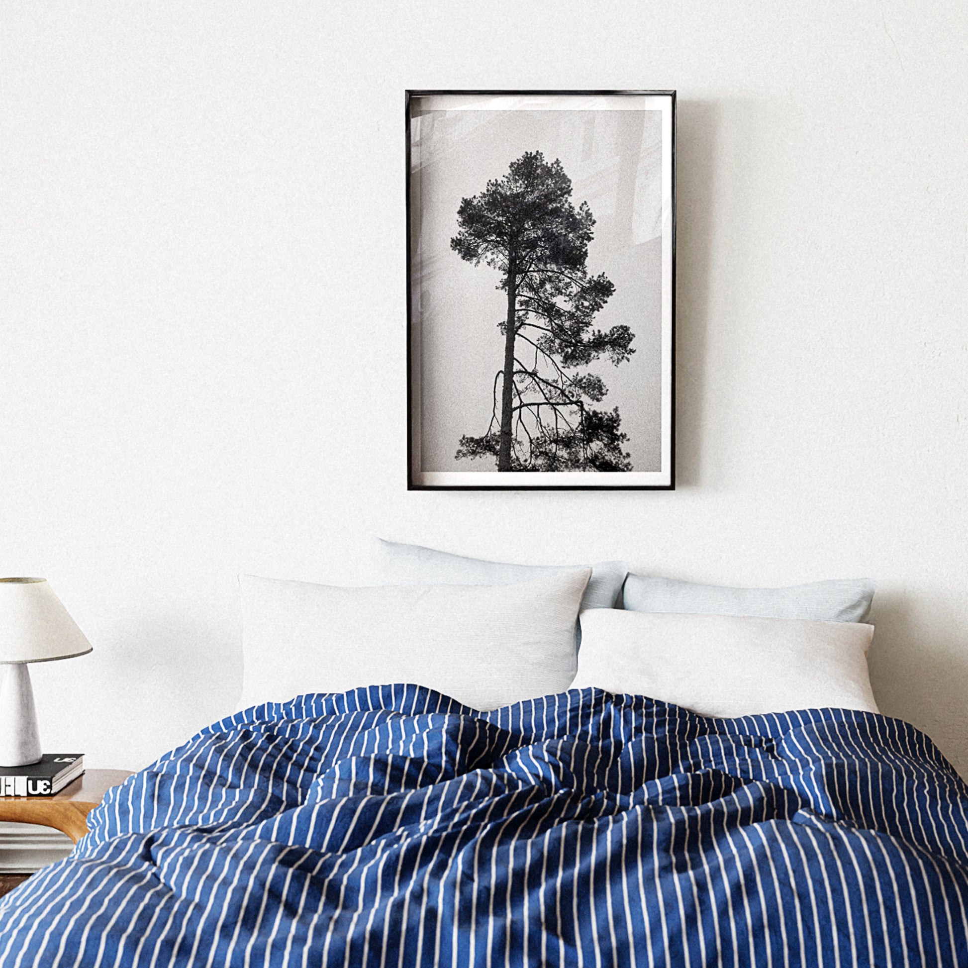 Framed black and white tree print on a wall above a bed with blue and white striped bedding.