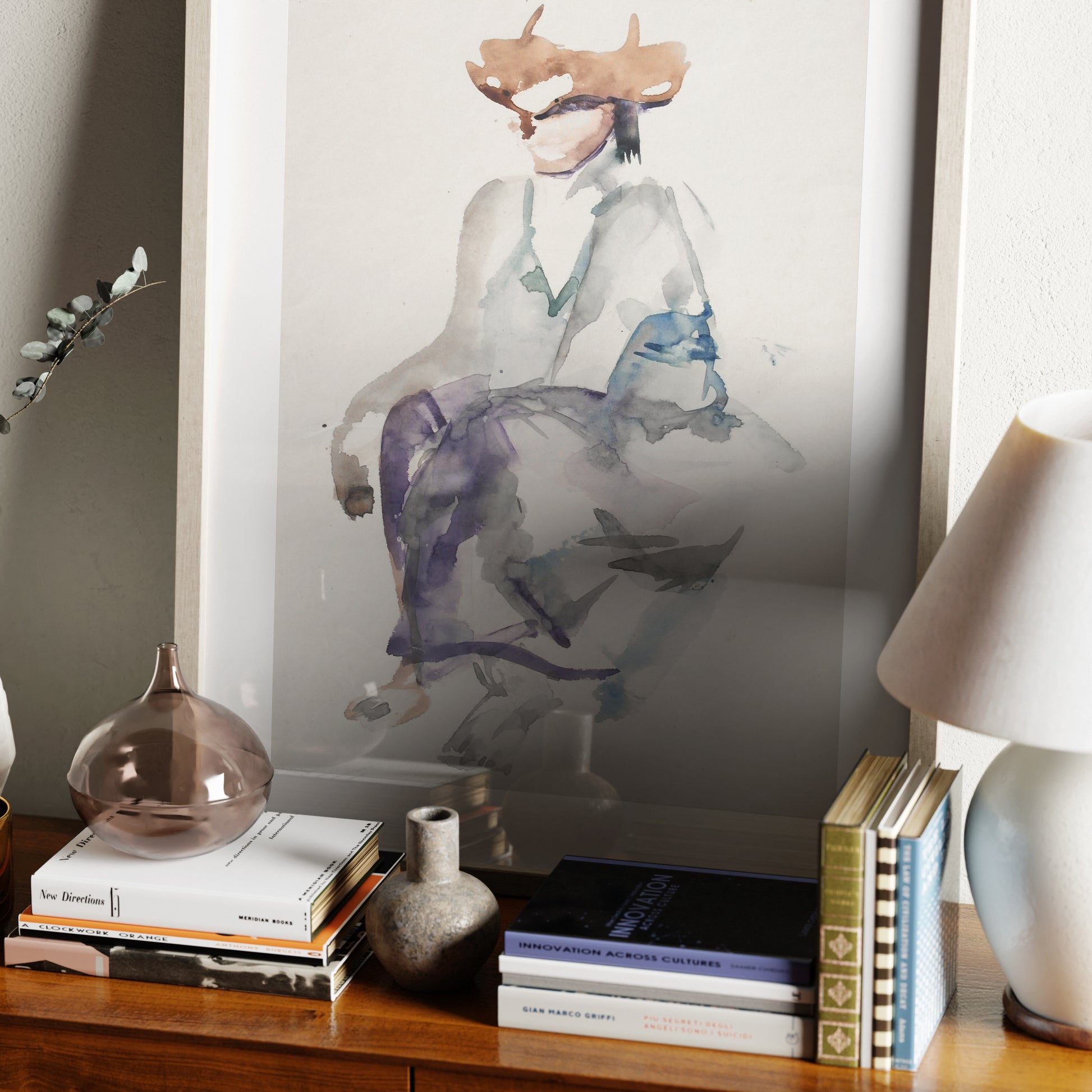 Naive watercolor figure study art print on a sideboard with books.